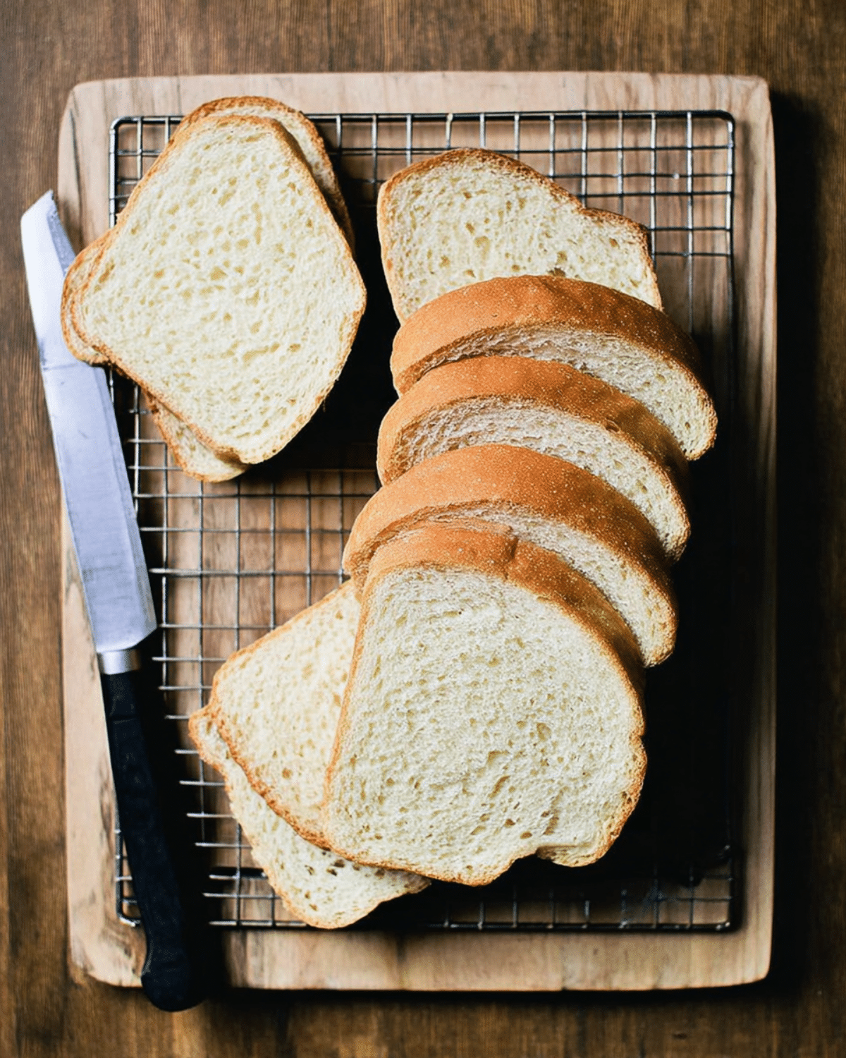Slices of soft sourdough sandwich bread on a cooling rack with a bread knife on a wooden surface.