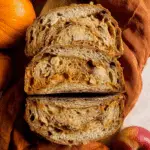 Sliced sourdough pumpkin apple bread with a golden crust and swirls of pumpkin and apple chunks, surrounded by pumpkins and apples.