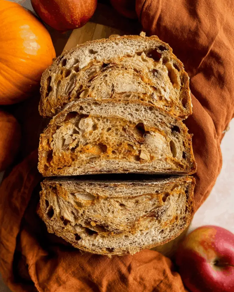 Sliced sourdough pumpkin apple bread with a golden crust and swirls of pumpkin and apple chunks, surrounded by pumpkins and apples.