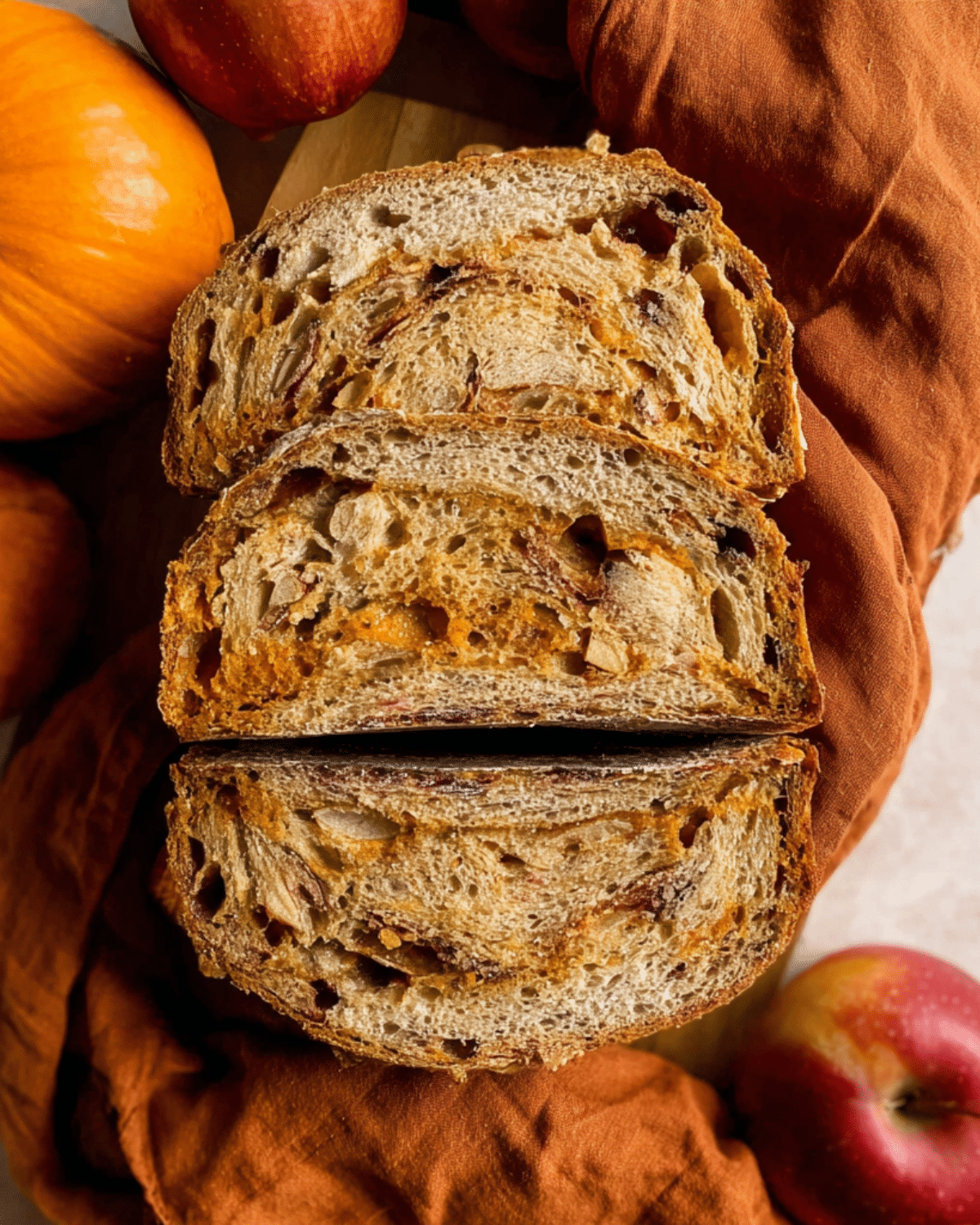 Sliced sourdough pumpkin apple bread with a golden crust and swirls of pumpkin and apple chunks, surrounded by pumpkins and apples.