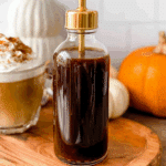Glass bottle filled with homemade Starbucks pumpkin spice coffee syrup on a wooden board with a pumpkin spice latte and pumpkins in the background.