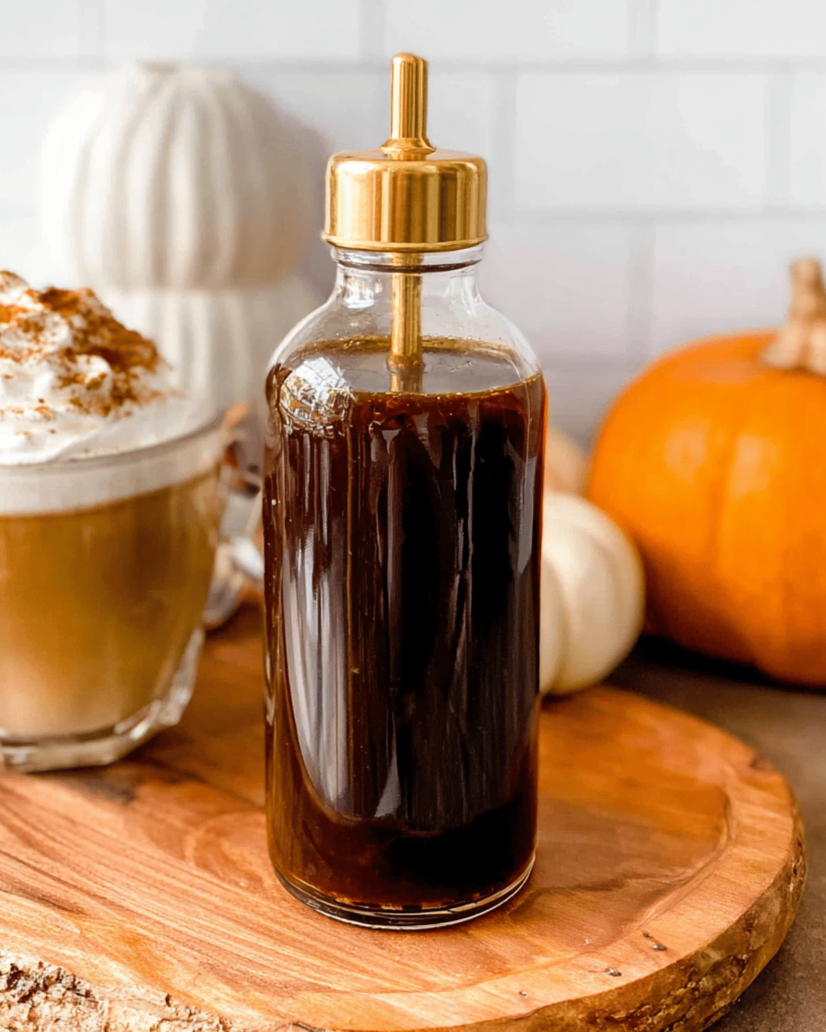 Glass bottle filled with homemade Starbucks pumpkin spice coffee syrup on a wooden board with a pumpkin spice latte and pumpkins in the background.