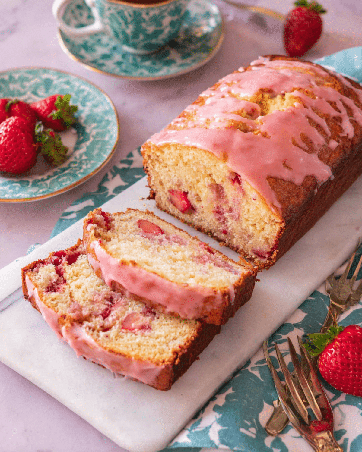 Sliced strawberry bread loaf with strawberry glaze on a marble board, surrounded by fresh strawberries and tea cups.