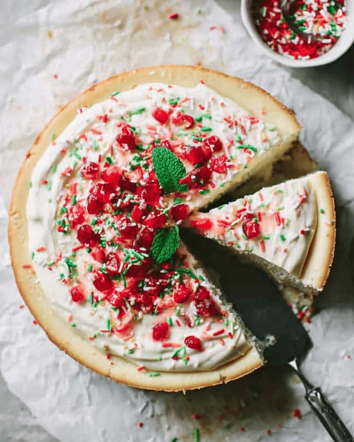 Sugar Cookie Cheesecake topped with whipped cream, festive sprinkles, red candy pieces, and fresh mint leaves.