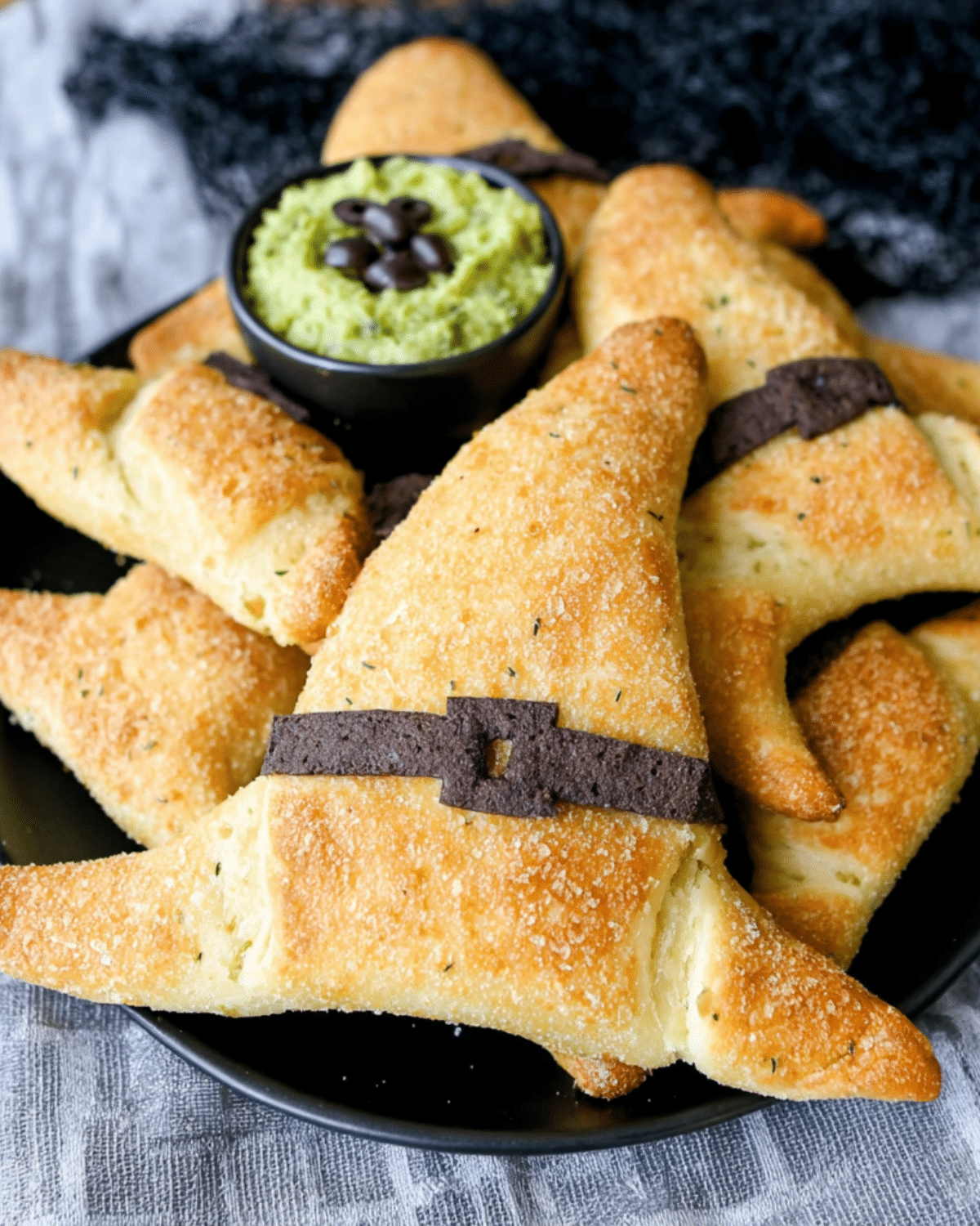 Witch Hat Crescent Rolls shaped like witch hats with black edible belts, served on a black plate with green dip garnished with black olives.