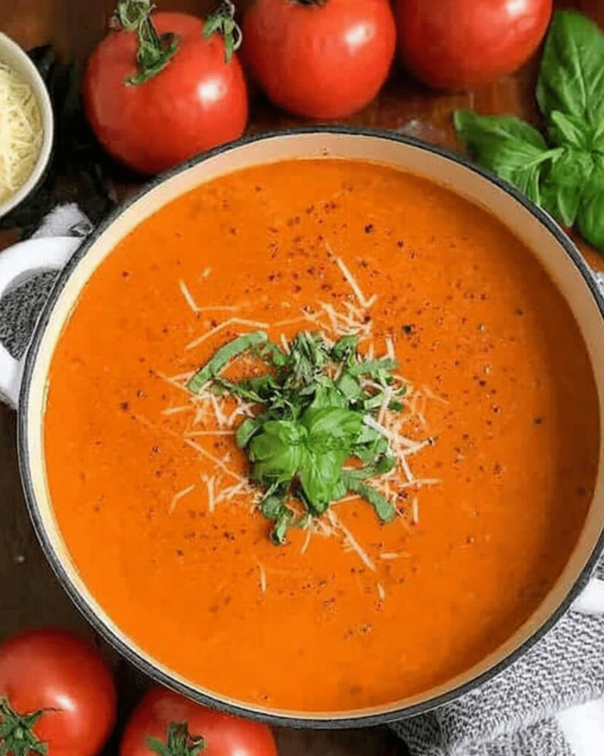 Tomato soup garnished with fresh basil and shredded cheese, surrounded by fresh tomatoes and basil leaves.