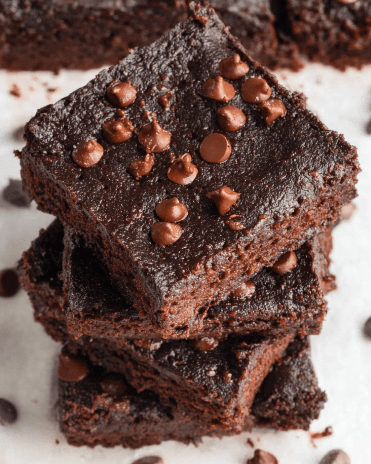 A stack of rich, fudgy 3 Ingredient Brownies topped with chocolate chips on a white parchment surface.