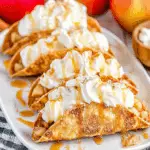 Crispy Apple Pie Tacos topped with whipped cream and caramel sauce, arranged on a white plate with fresh apples in the background.