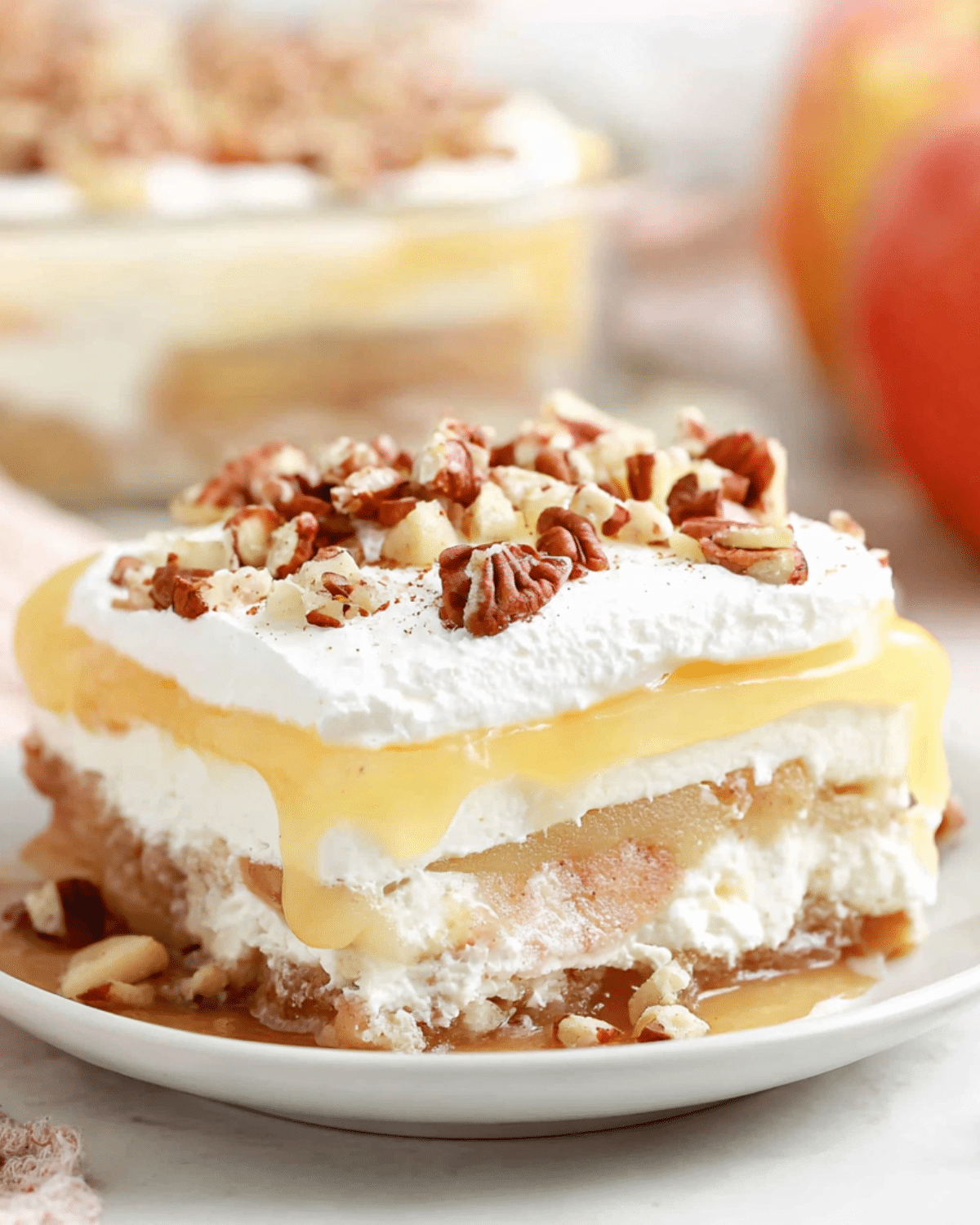 A slice of Apple Delight dessert with layers of whipped cream, apple filling, pudding, and a nutty crust topped with pecans.