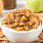 A white bowl filled with warm Apple Pie Dip topped with tender cinnamon-spiced apple chunks, surrounded by cinnamon sugar chips.