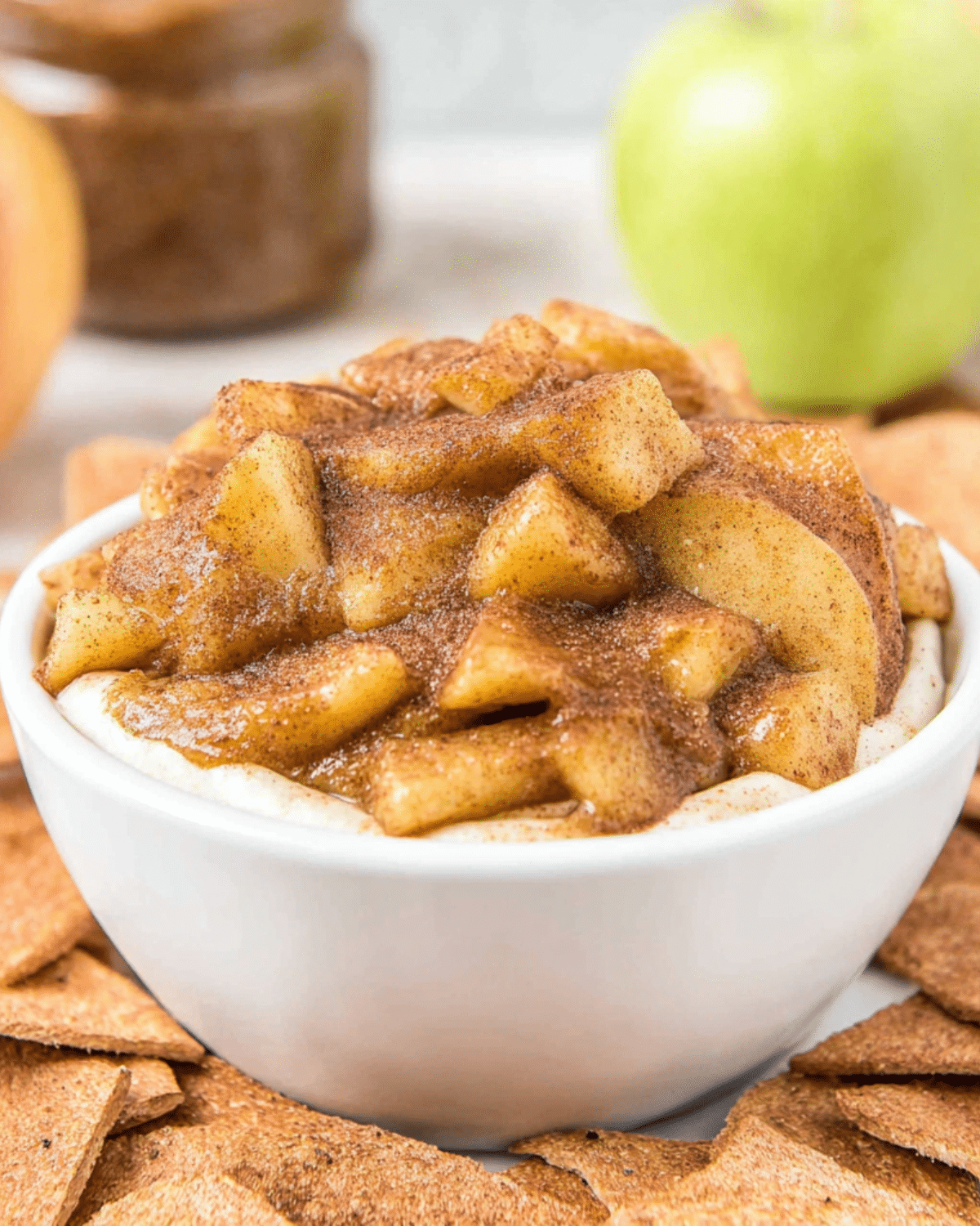 A white bowl filled with warm Apple Pie Dip topped with tender cinnamon-spiced apple chunks, surrounded by cinnamon sugar chips.