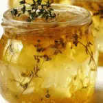 A glass jar of apple thyme jam filled with golden jelly, apple chunks, and fresh thyme sprigs.