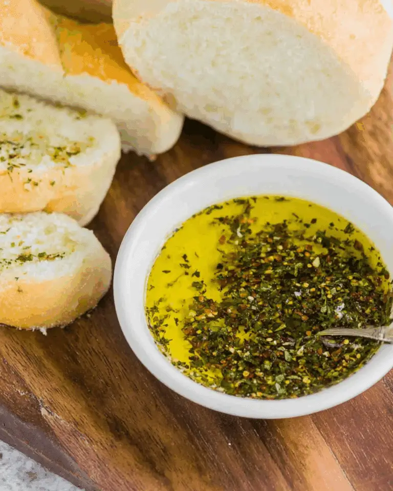 A bowl of bread dipping oil with herbs and spices, served alongside sliced bread on a wooden board.