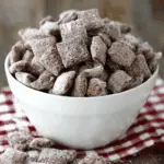 A white bowl filled with brownie batter muddy buddies coated in powdered sugar, with pieces scattered on a red checkered cloth.