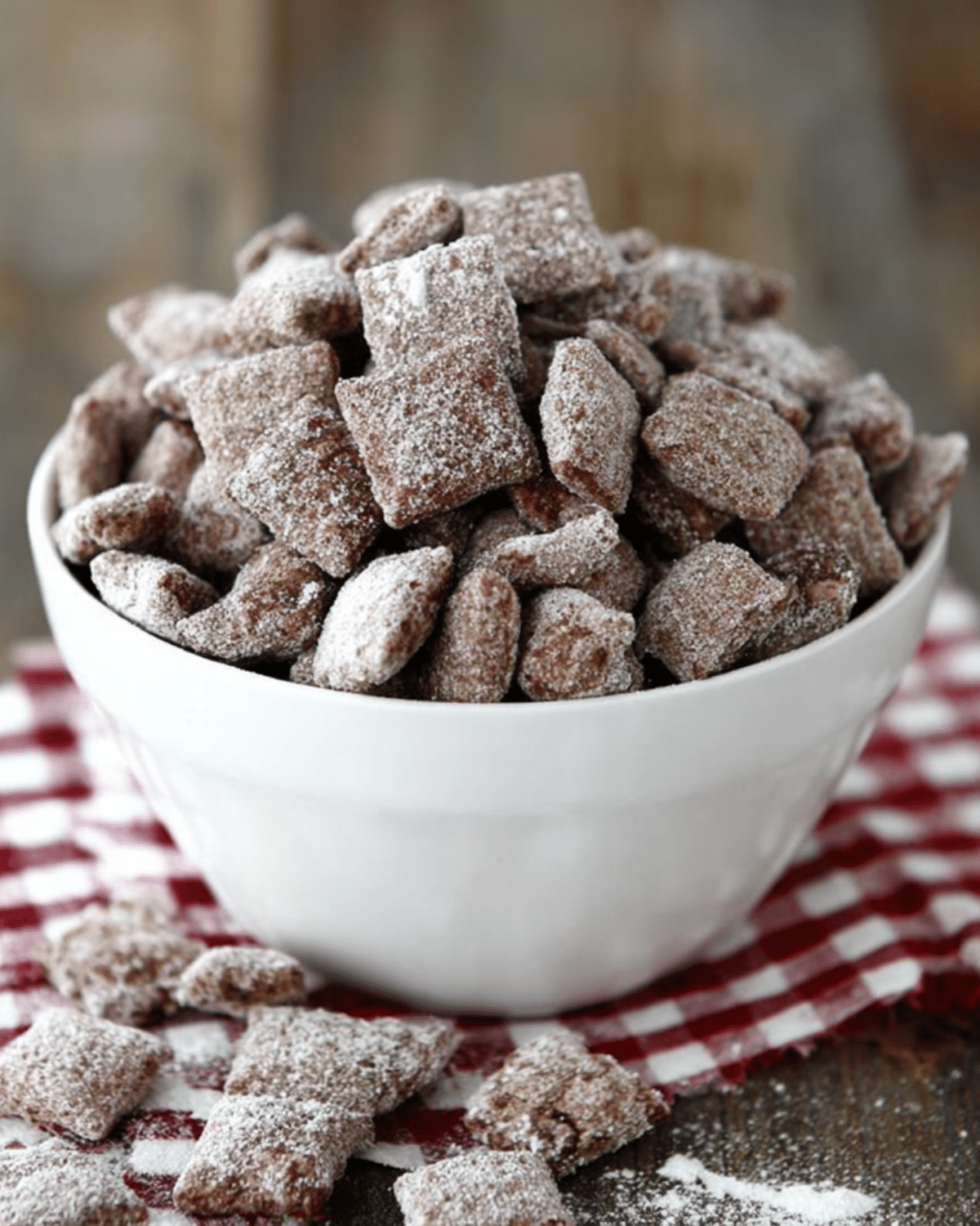 A white bowl filled with brownie batter muddy buddies coated in powdered sugar, with pieces scattered on a red checkered cloth.