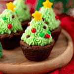 Festive Brownie Cups Christmas Tree desserts topped with green frosting, candy ornaments, and yellow star toppers, served on a wooden plate.