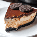 Slice of BUCKEYE PIE with a chocolate cookie crust, creamy peanut butter filling, rich chocolate topping, and Oreo cookies on top.