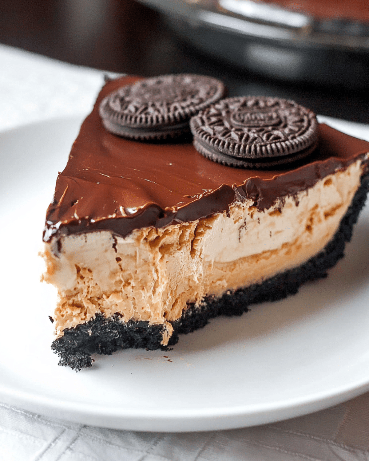 Slice of BUCKEYE PIE with a chocolate cookie crust, creamy peanut butter filling, rich chocolate topping, and Oreo cookies on top.