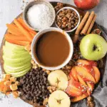 A festive Caramel Apple Dip Board with sliced apples, chocolate chips, caramel candies, nuts, shredded coconut, and a bowl of caramel dip.