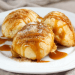 Three caramel apple pie bombs on a white plate, dusted with cinnamon sugar and drizzled with caramel sauce.