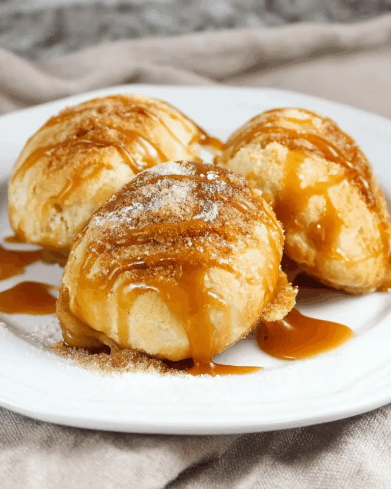 Three caramel apple pie bombs on a white plate, dusted with cinnamon sugar and drizzled with caramel sauce.