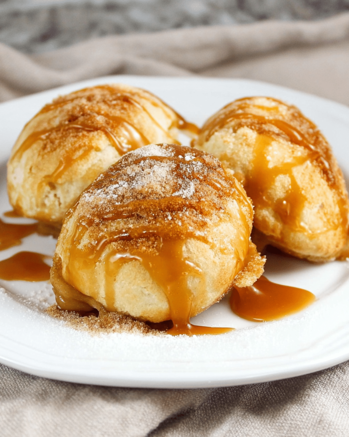 Three caramel apple pie bombs on a white plate, dusted with cinnamon sugar and drizzled with caramel sauce.