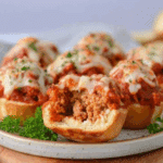 A plate of Cheesy Meatball Bombs topped with melted mozzarella and parsley, with one cut open to reveal the juicy meatball filling.