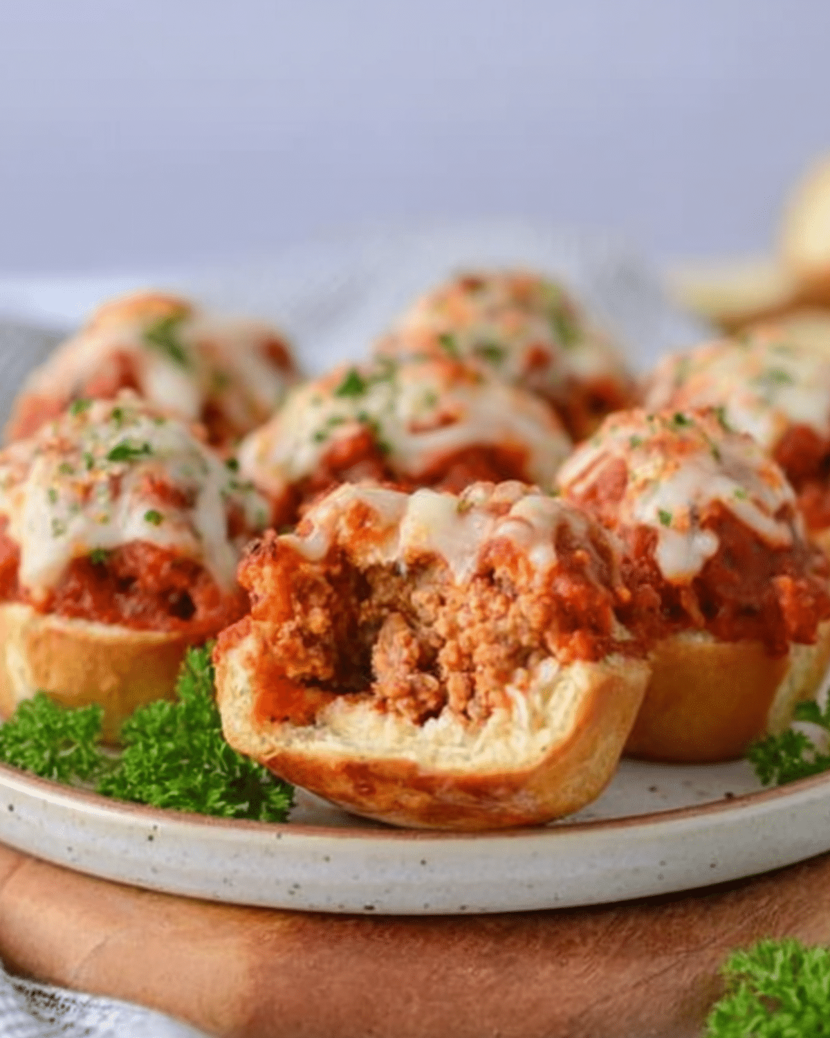 A plate of Cheesy Meatball Bombs topped with melted mozzarella and parsley, with one cut open to reveal the juicy meatball filling.