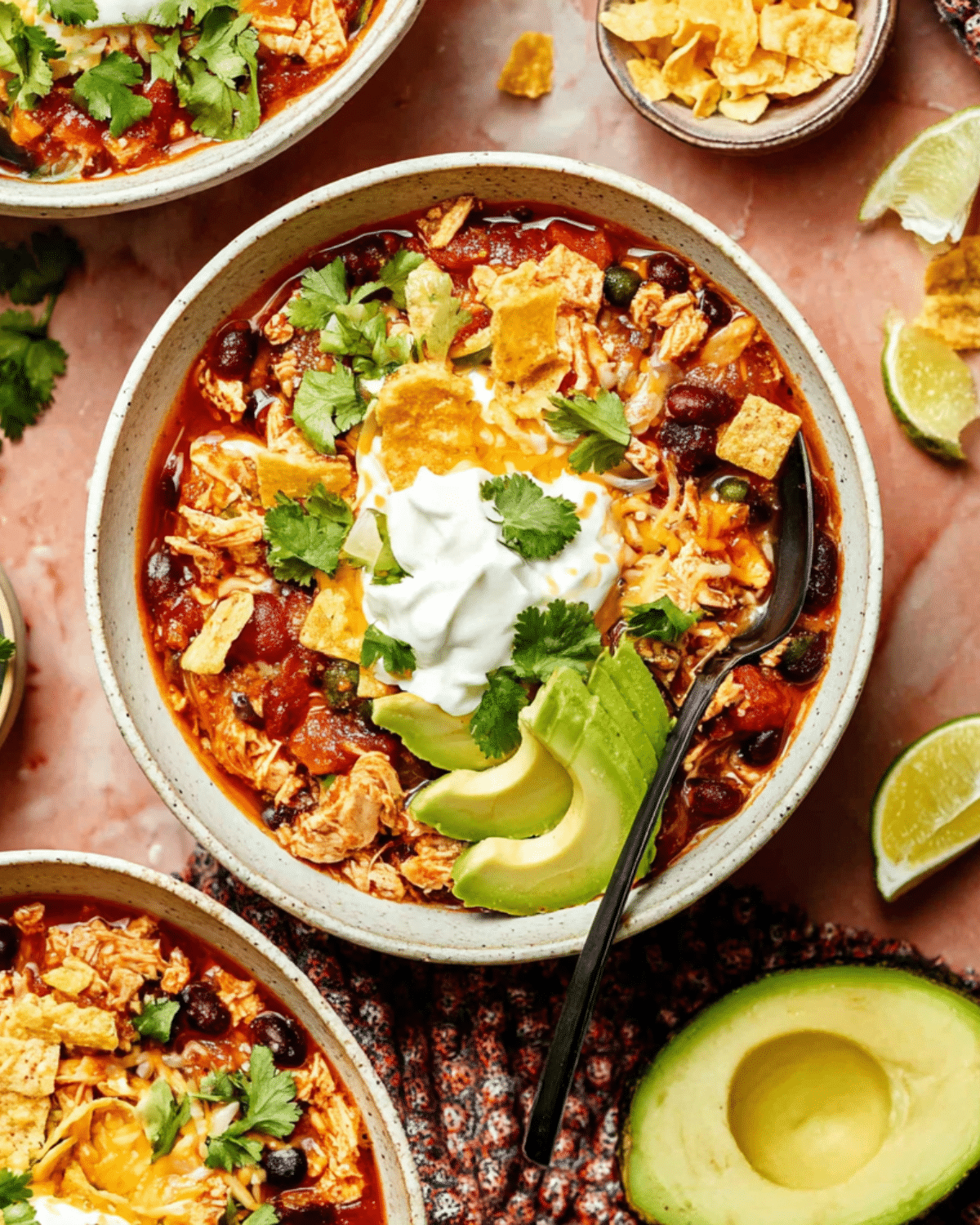 A bowl of Chicken Enchilada Chili topped with sour cream, sliced avocado, shredded cheese, tortilla chips, and fresh cilantro.