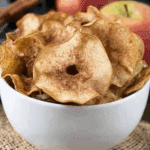 A white bowl filled with crispy Cinnamon Sugar Apple Chips, surrounded by fresh apples and a cinnamon stick.