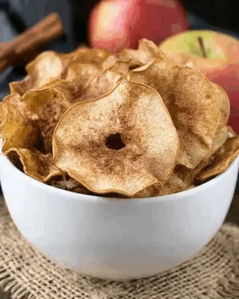 A white bowl filled with crispy Cinnamon Sugar Apple Chips, surrounded by fresh apples and a cinnamon stick.
