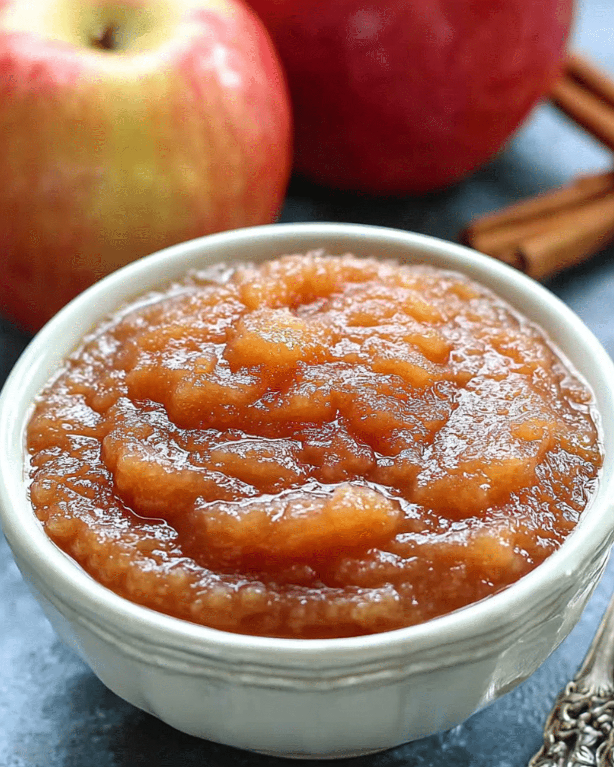 White bowl filled with Crockpot Cinnamon Applesauce, surrounded by fresh apples and cinnamon sticks.