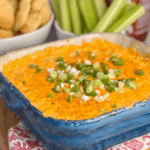 Frank’s Red Hot Buffalo Chicken Dip in a blue baking dish, topped with melted cheese, green onions, and crumbled cheese, served with celery sticks and bread.