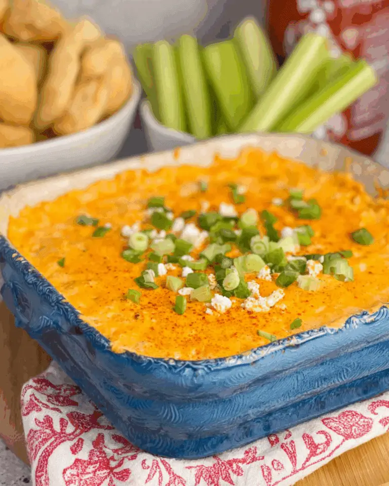 Frank’s Red Hot Buffalo Chicken Dip in a blue baking dish, topped with melted cheese, green onions, and crumbled cheese, served with celery sticks and bread.