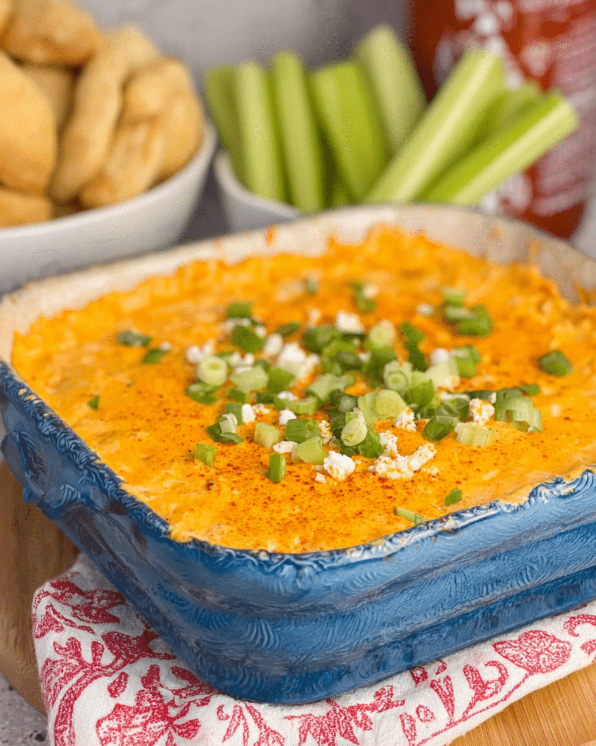 Frank’s Red Hot Buffalo Chicken Dip in a blue baking dish, topped with melted cheese, green onions, and crumbled cheese, served with celery sticks and bread.