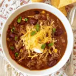 Ground Beef Stovetop Chili in a white bowl topped with shredded cheddar cheese, sour cream, and chopped green onions, served with tortilla chips.