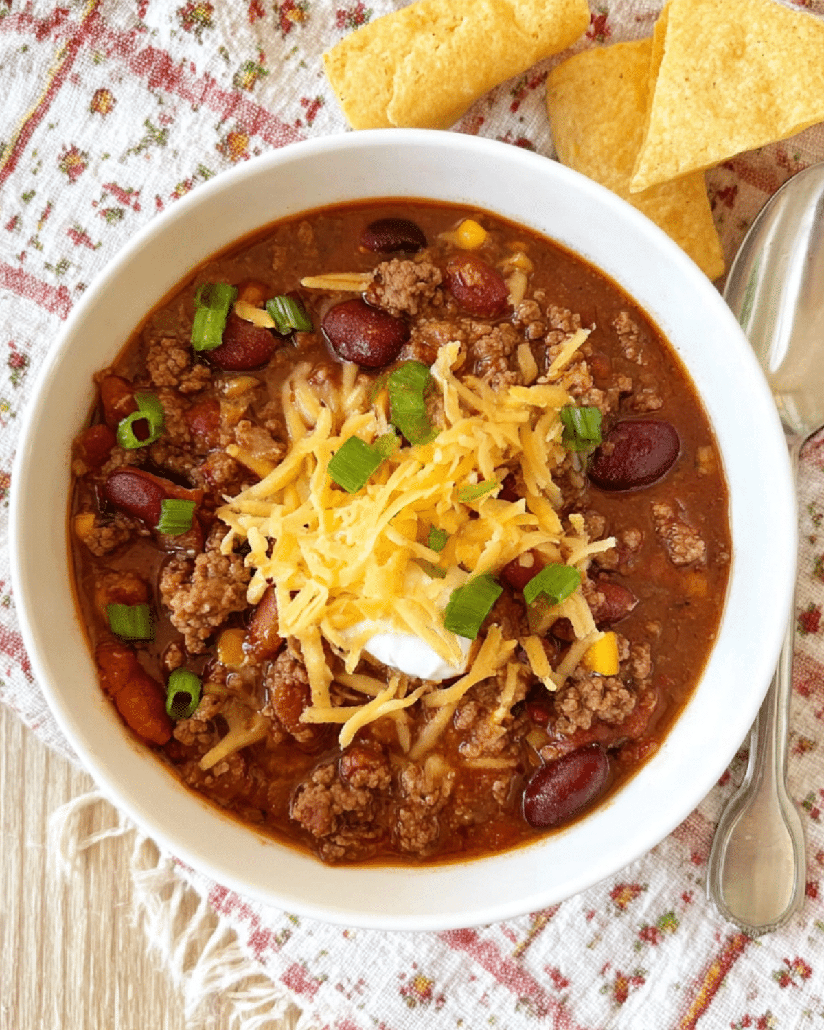 Ground Beef Stovetop Chili in a white bowl topped with shredded cheddar cheese, sour cream, and chopped green onions, served with tortilla chips.