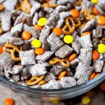 A glass bowl filled with Halloween Puppy Chow made with powdered sugar-coated cereal, pretzels, chocolate chips, and orange and yellow candies.