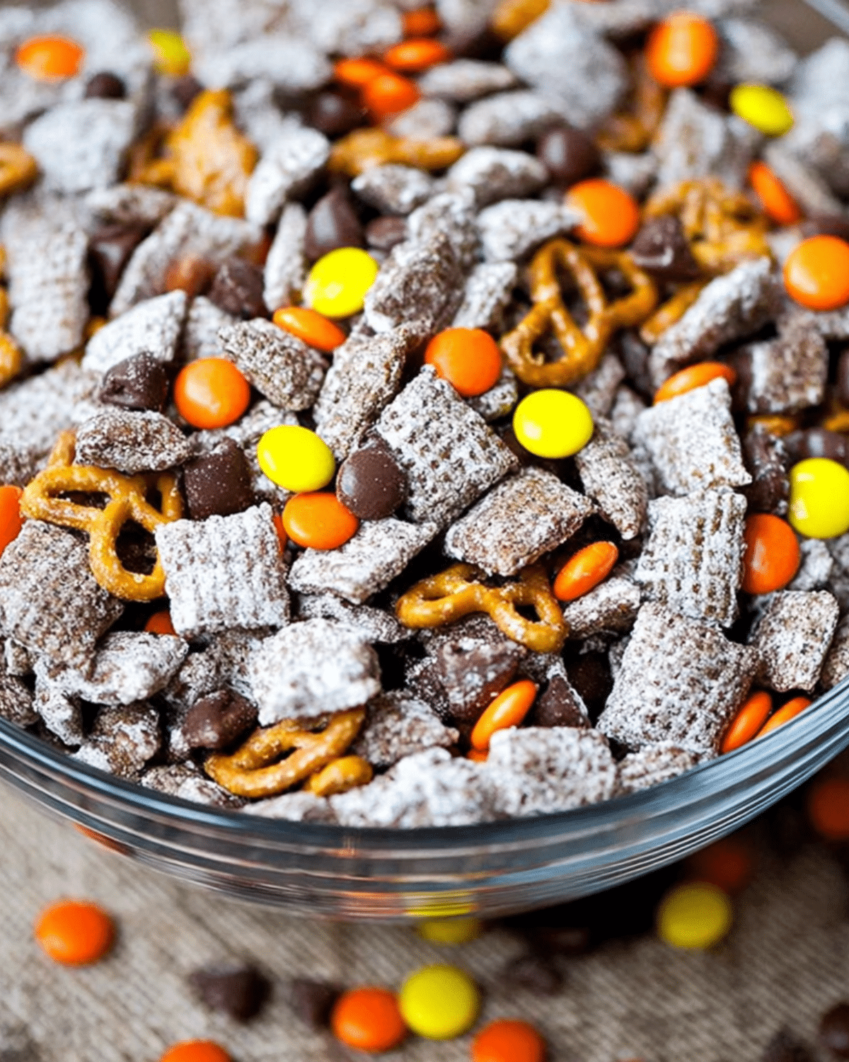 A glass bowl filled with Halloween Puppy Chow made with powdered sugar-coated cereal, pretzels, chocolate chips, and orange and yellow candies.