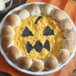 A festive dish of Halloween buffalo chicken dip decorated with a jack-o’-lantern face, surrounded by pull-apart bread rolls.