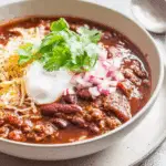 Homemade Ground Beef Chili served in a bowl with red kidney beans, topped with shredded cheese, sour cream, chopped red onions, and fresh cilantro.