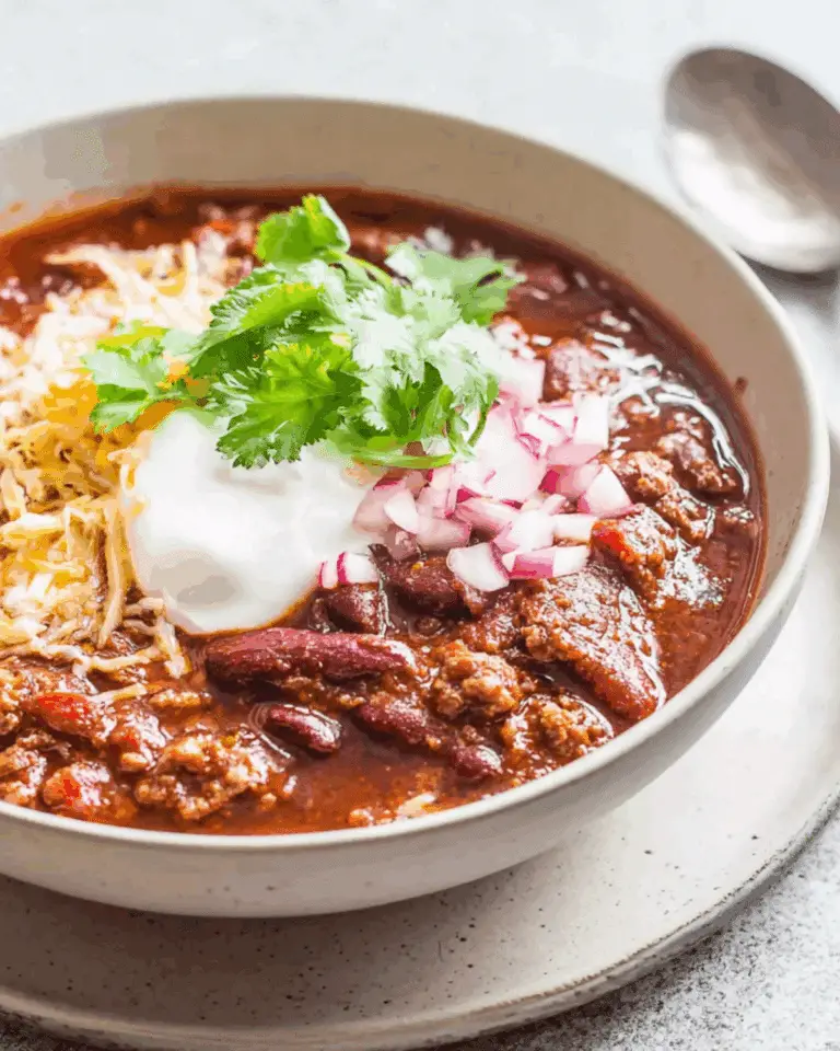 Homemade Ground Beef Chili served in a bowl with red kidney beans, topped with shredded cheese, sour cream, chopped red onions, and fresh cilantro.