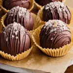 Hot chocolate bombs coated in dark chocolate, drizzled with chocolate, and sprinkled with sugar crystals, displayed in paper wrappers.
