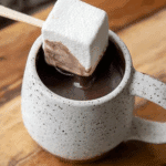 Hot chocolate on a stick with a large square marshmallow dipped into a mug of rich, creamy hot cocoa.