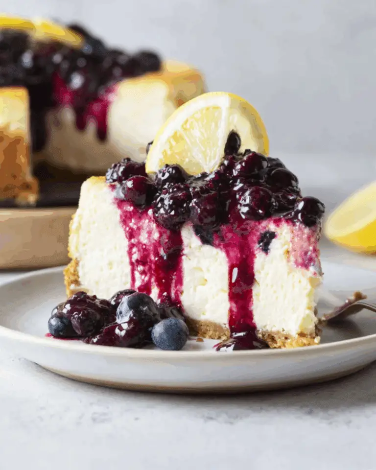 A slice of lemon blueberry cheesecake topped with blueberry compote and a fresh lemon wedge, served on a white plate.