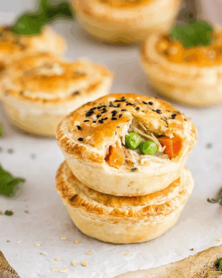 Golden-brown mini chicken pot pies stacked on parchment paper, with a flaky crust and visible filling of peas, carrots, and shredded chicken.