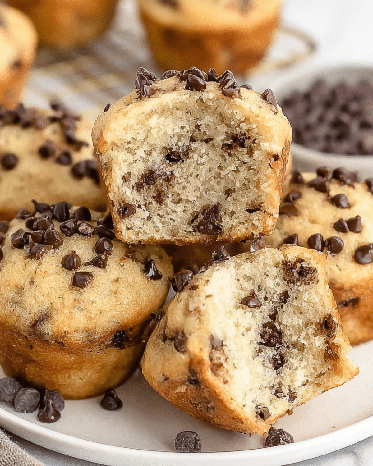 A plate of Mini Chocolate Chip Muffins, with one muffin cut open to reveal its soft, fluffy texture and chocolate chips inside.