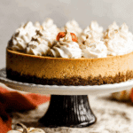 A No Bake Pumpkin Cheesecake topped with whipped cream swirls and a mini decorative pumpkin, displayed on a cake stand.
