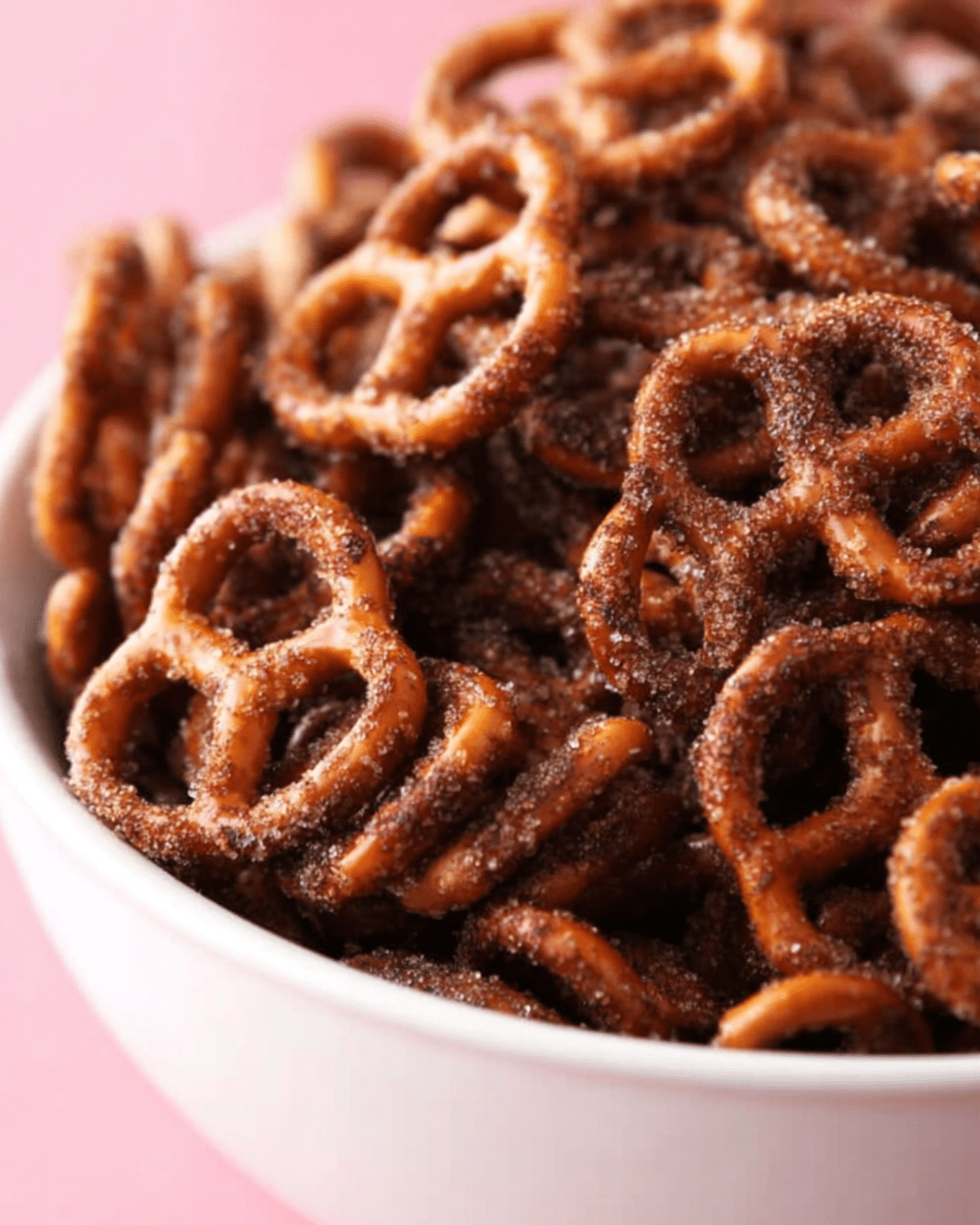 Close-up of a bowl filled with Oven Cinnamon Pretzels coated in a rich cinnamon-sugar mixture against a pink background.