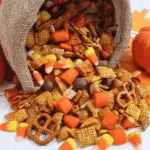 A festive assortment of Pumpkin Spice Snack Mix spilling out of a burlap-wrapped container, featuring candy corn, cereal squares, pretzels, and fall-themed sweets.